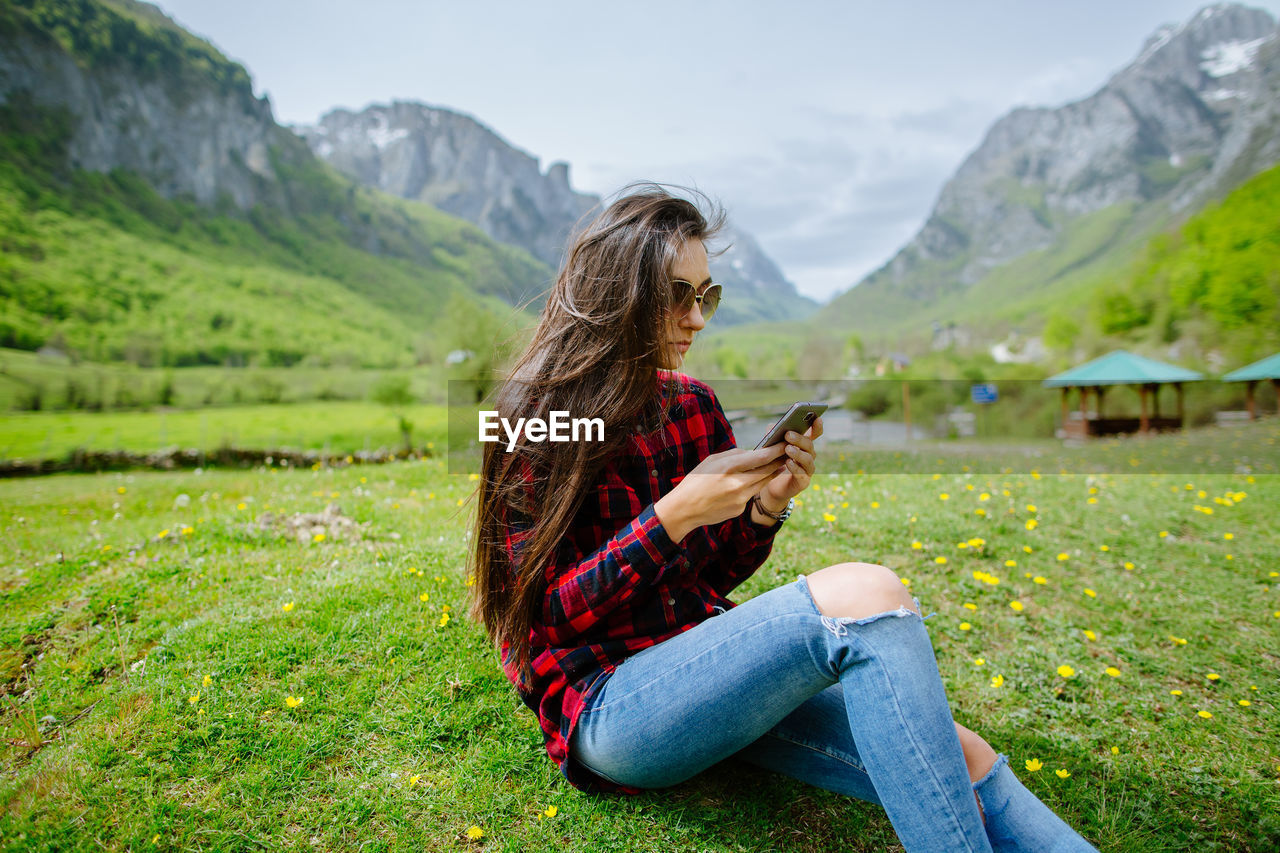 Woman using phone while sitting on grassy field against mountains