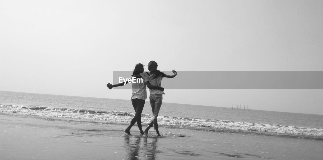 Full length rear view of friends walking on shore at beach against clear sky