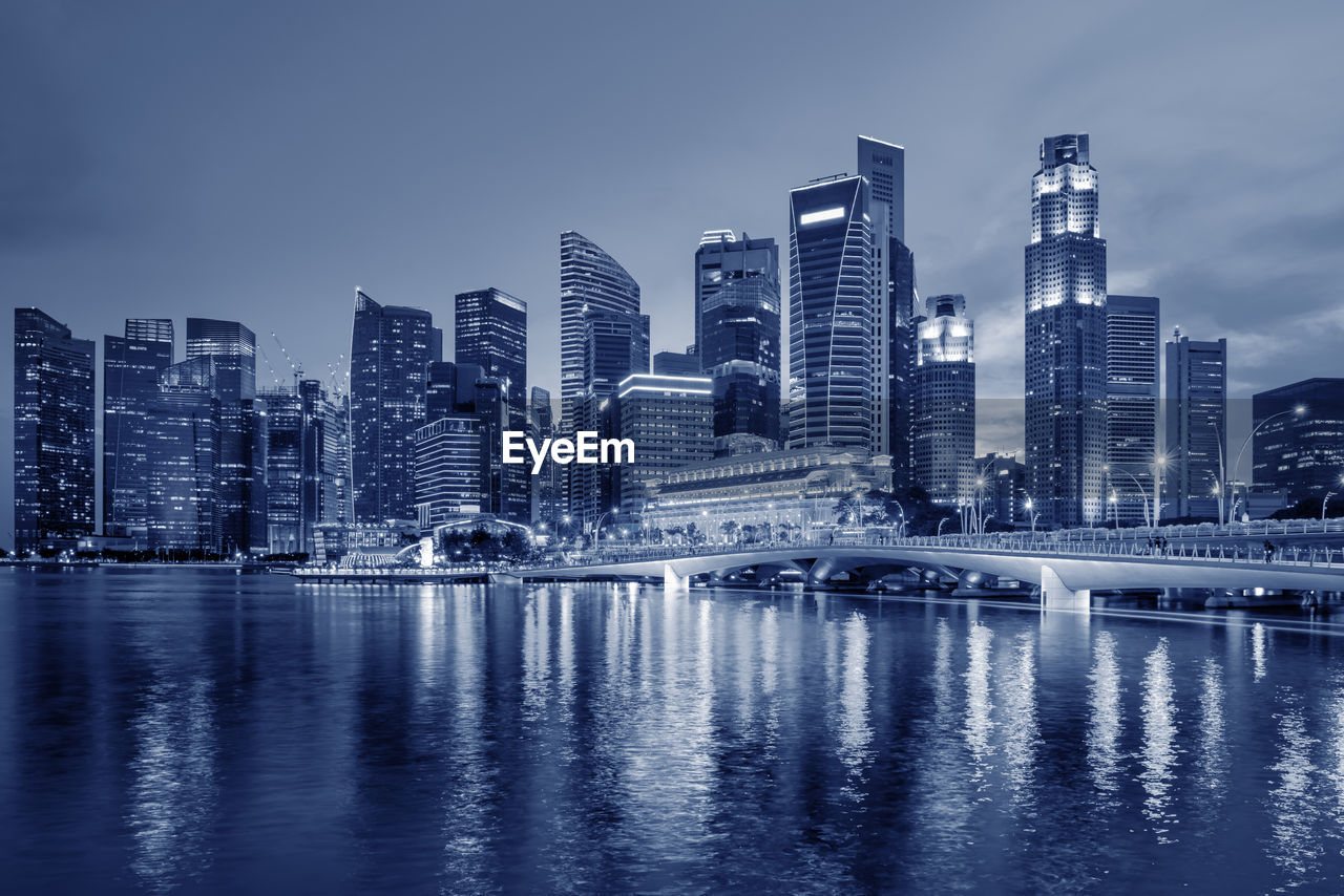 Illuminated city by river against sky at dusk