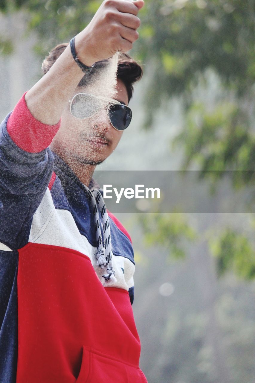 Portrait of young man wearing sunglasses throwing sand outdoors