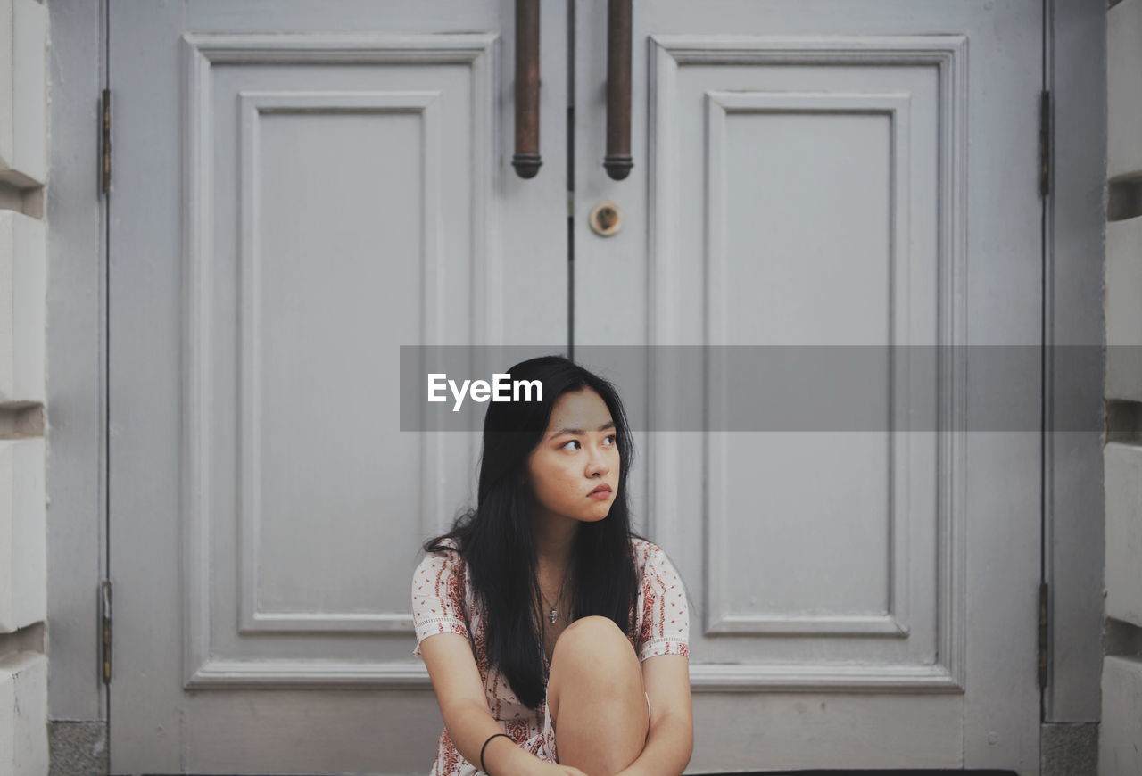 Young woman looking away while sitting against door