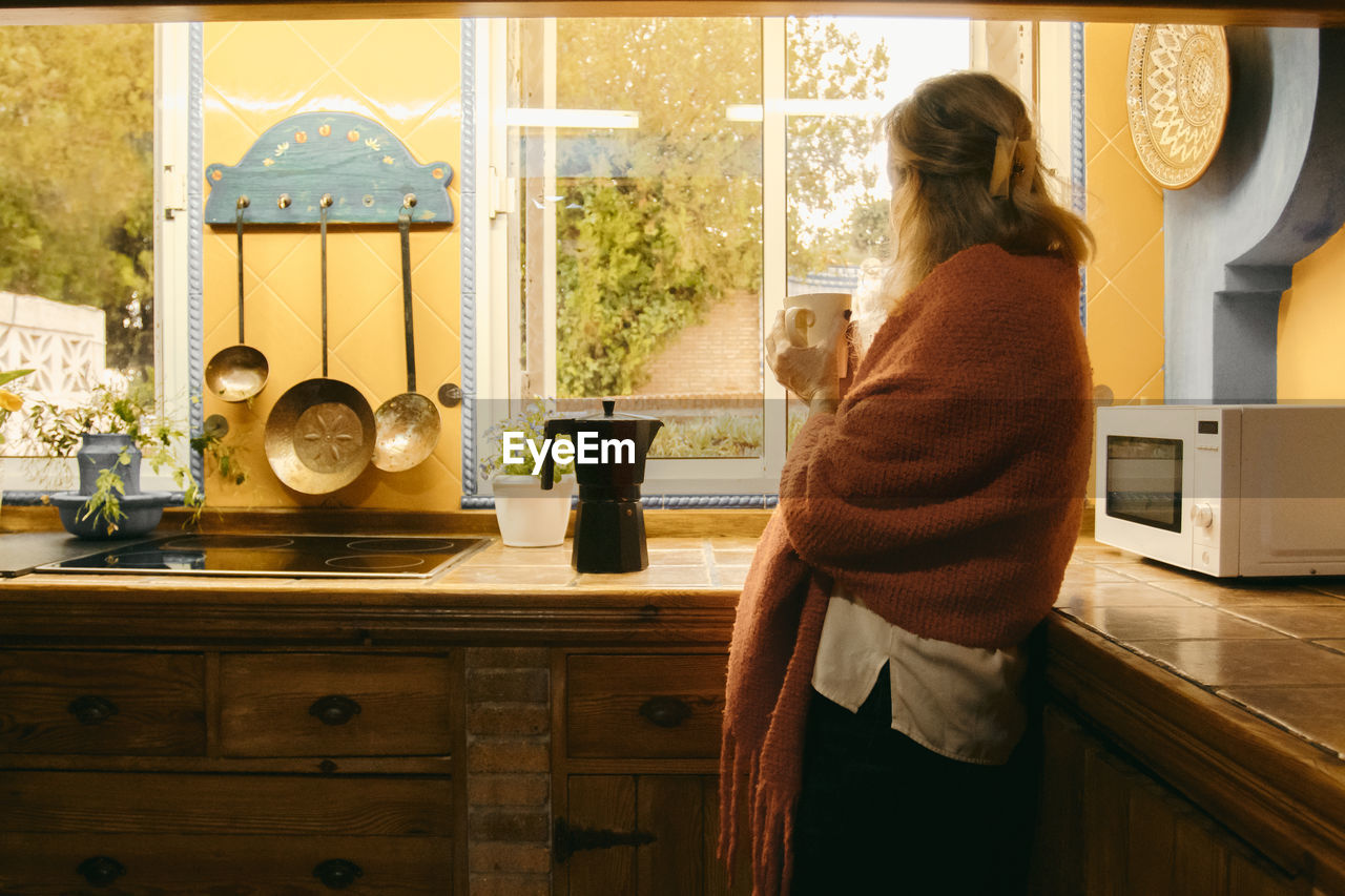 Senior woman in covered blanket looking through window while drinking coffee at home