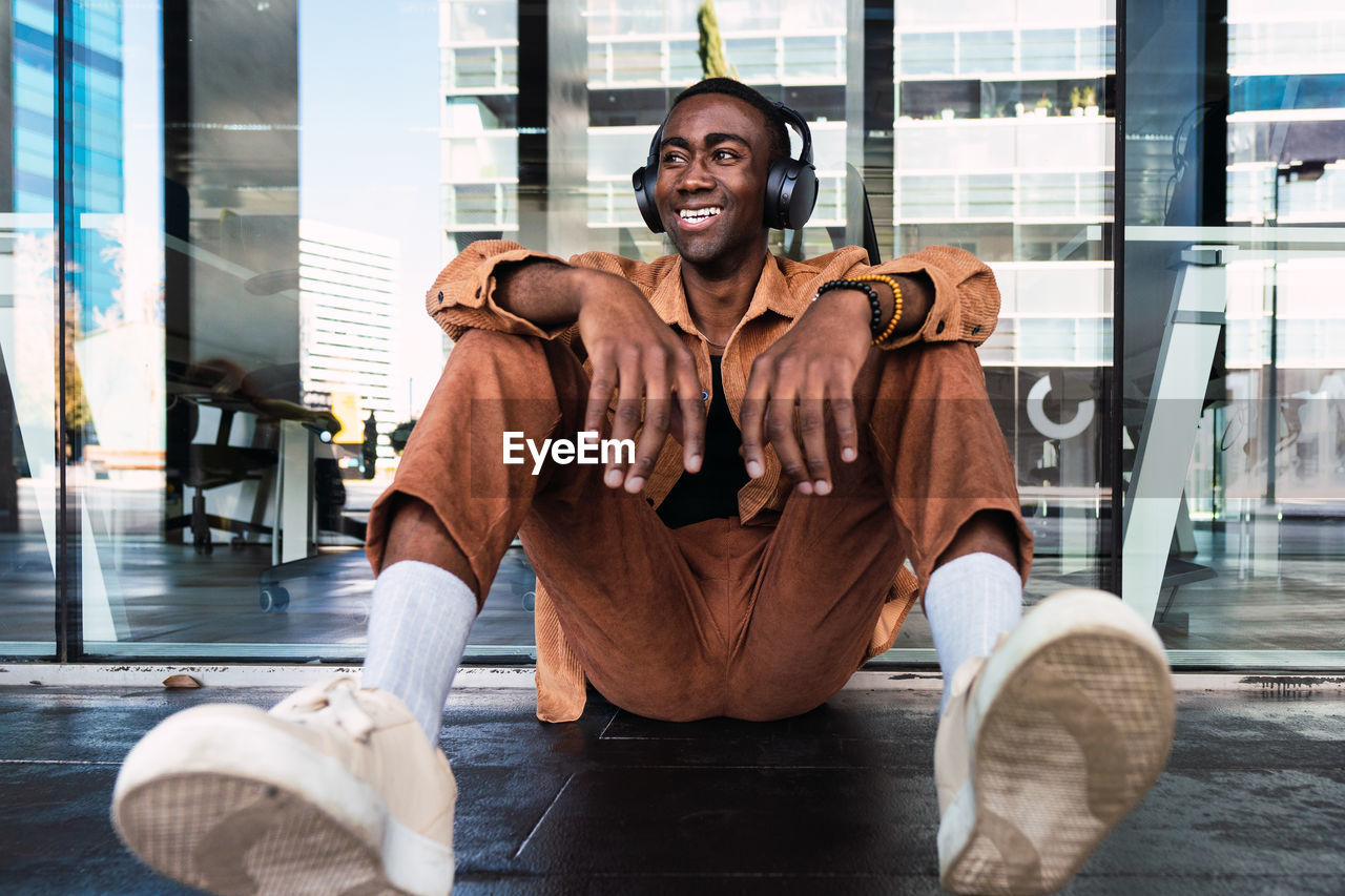 Stylish african american male listening to music in wireless headphones and looking away while sitting near glass wall in city