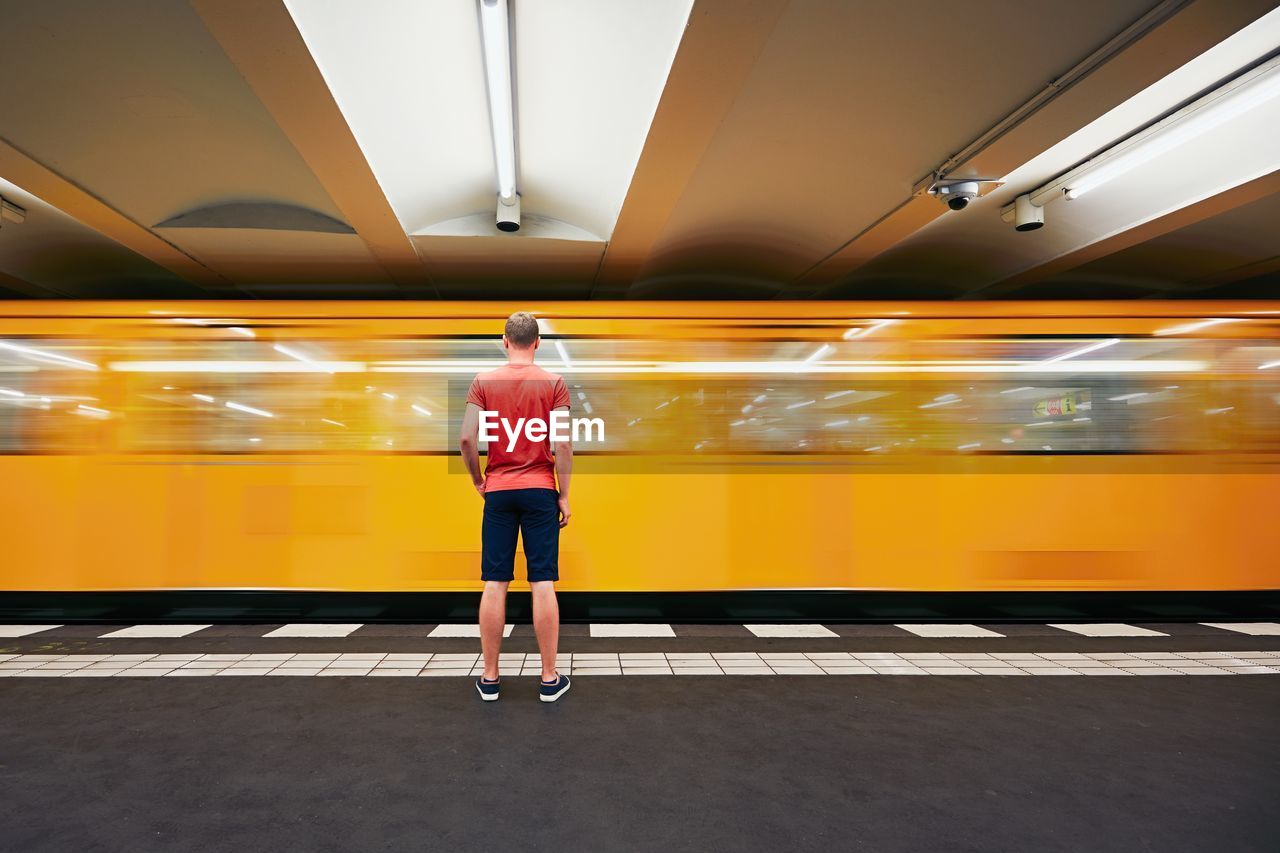 Rear view full length of man standing against blurred motion of yellow train at station
