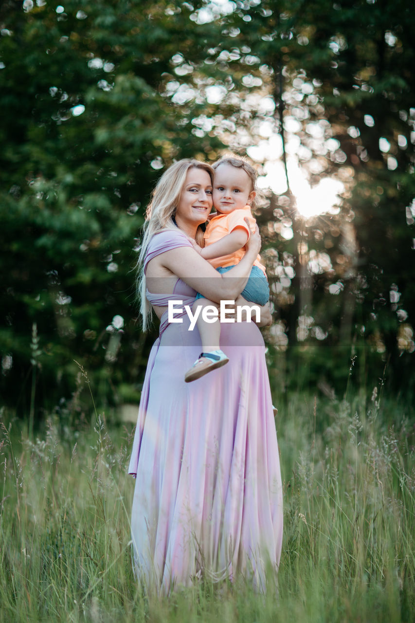 Happy pregnant woman carrying son while standing on field