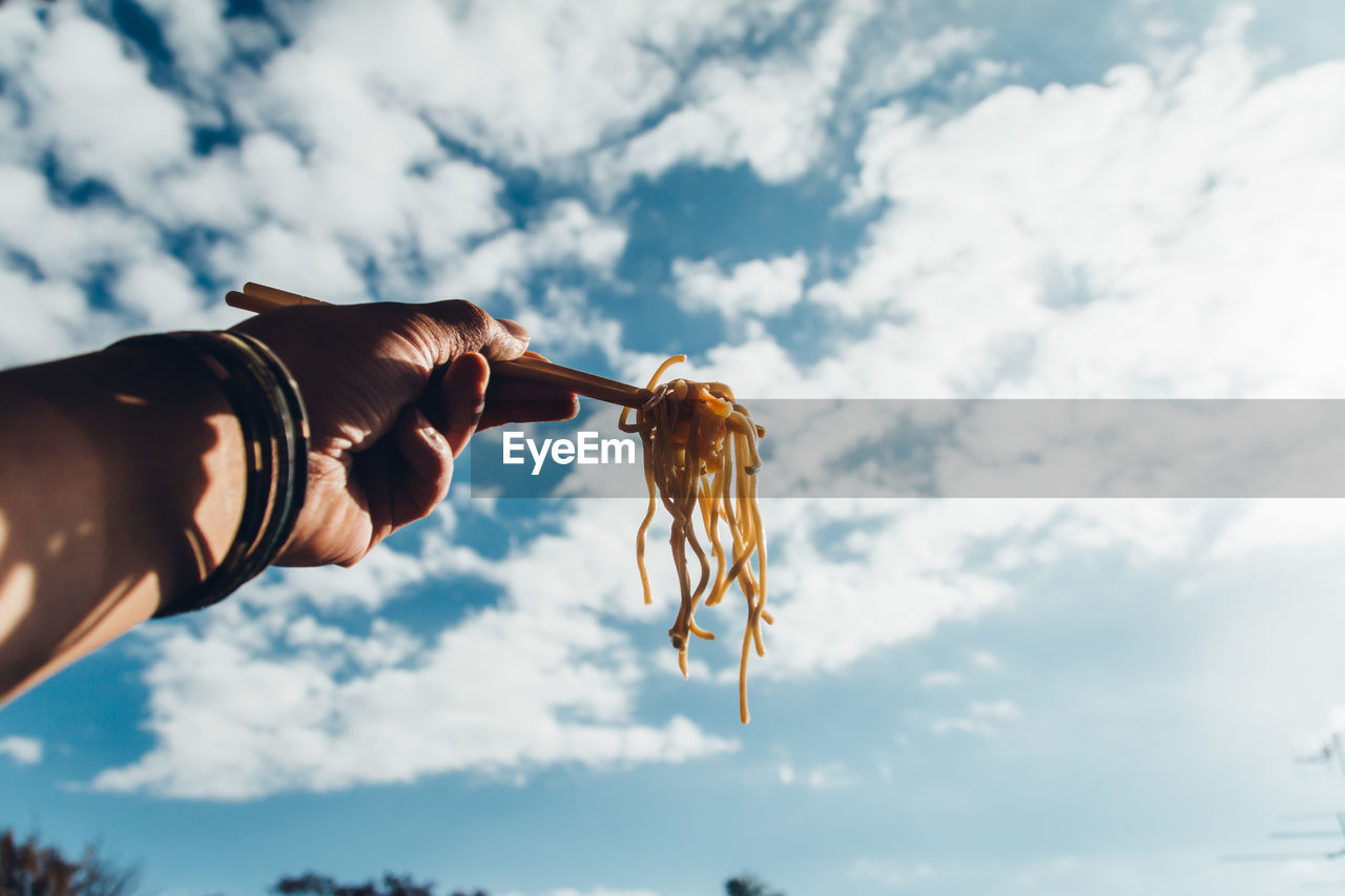 Cropped hand holding noodles against sky