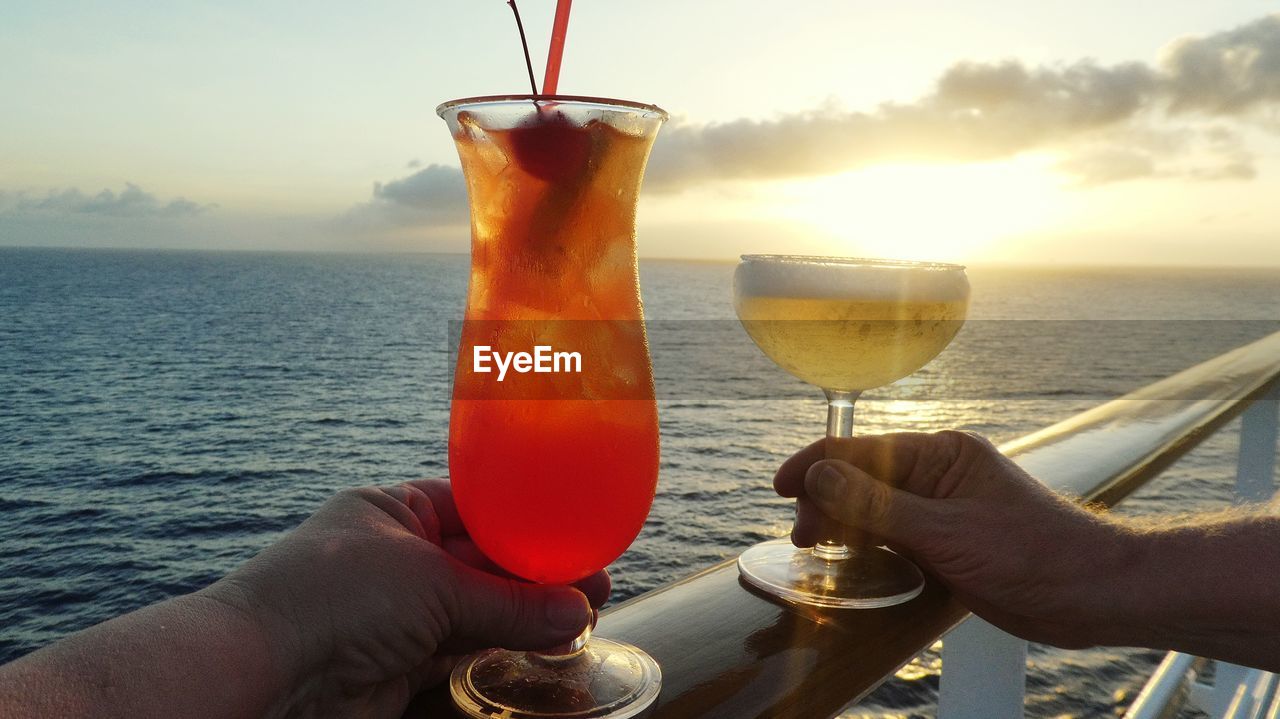 Close-up of hands holding cocktails against sea at sunset