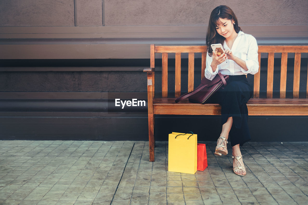 Woman using mobile phone while sitting on bench in city