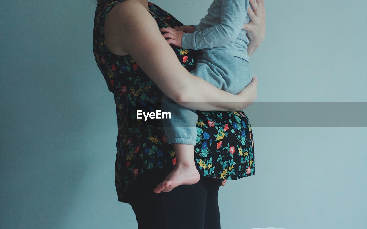 Cropped image of pregnant woman carrying son while standing against wall