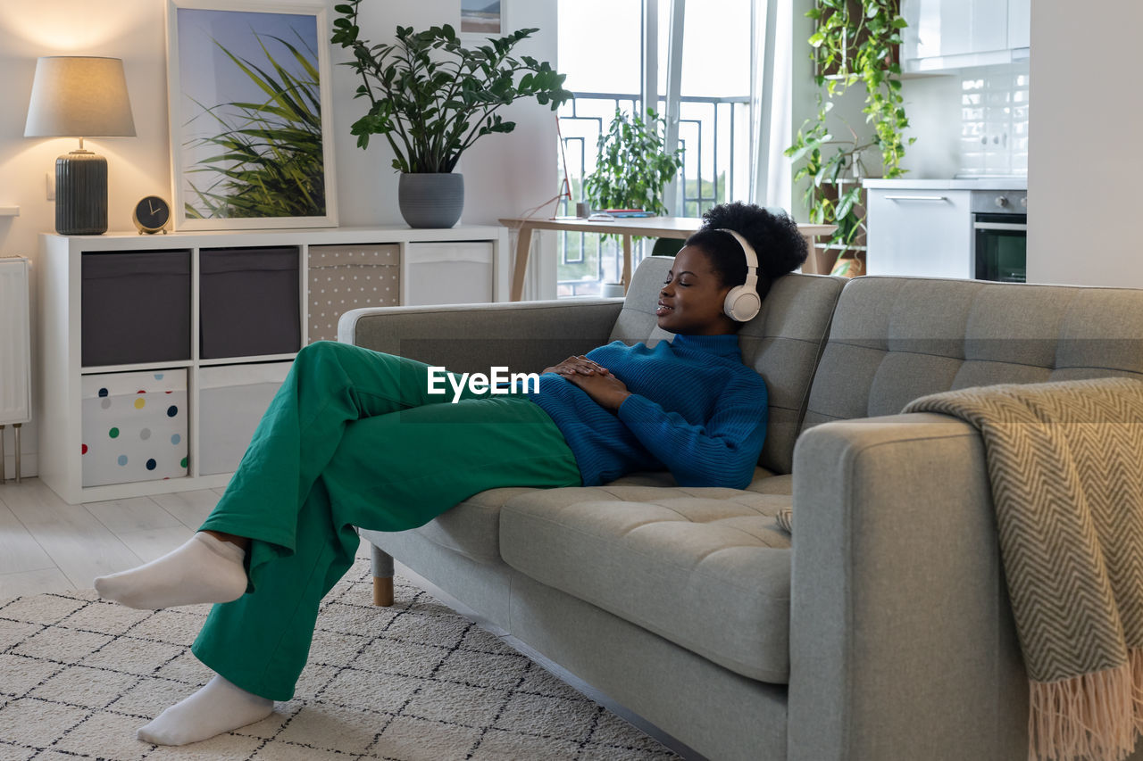 Happy relaxed african american woman in headphones closing eyes enjoying music sits on sofa