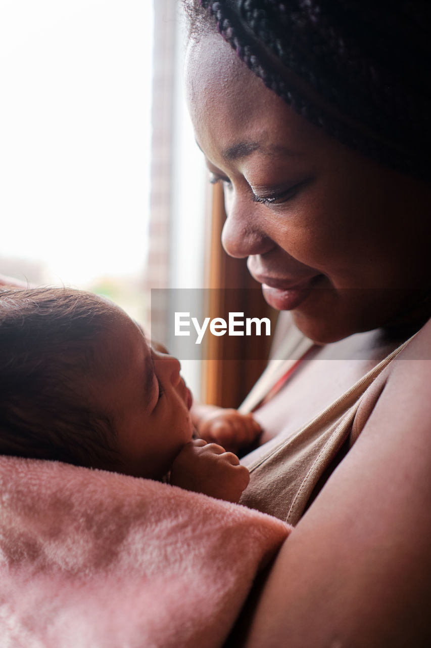 Side view of african american female with long hair standing at window and kissing newborn baby wrapped in soft towel