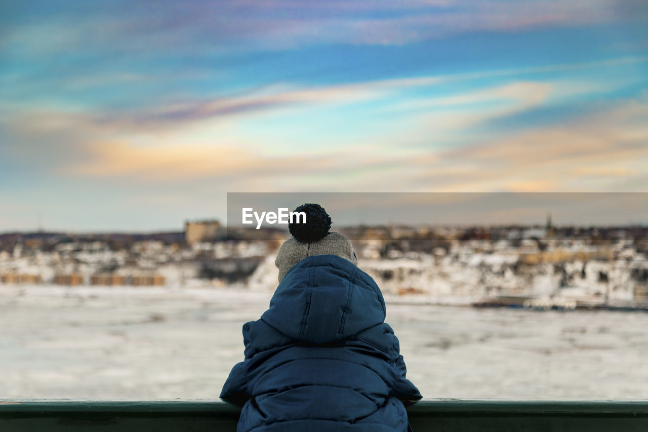 6 years old boy in winter clothes watching the sunset over the river