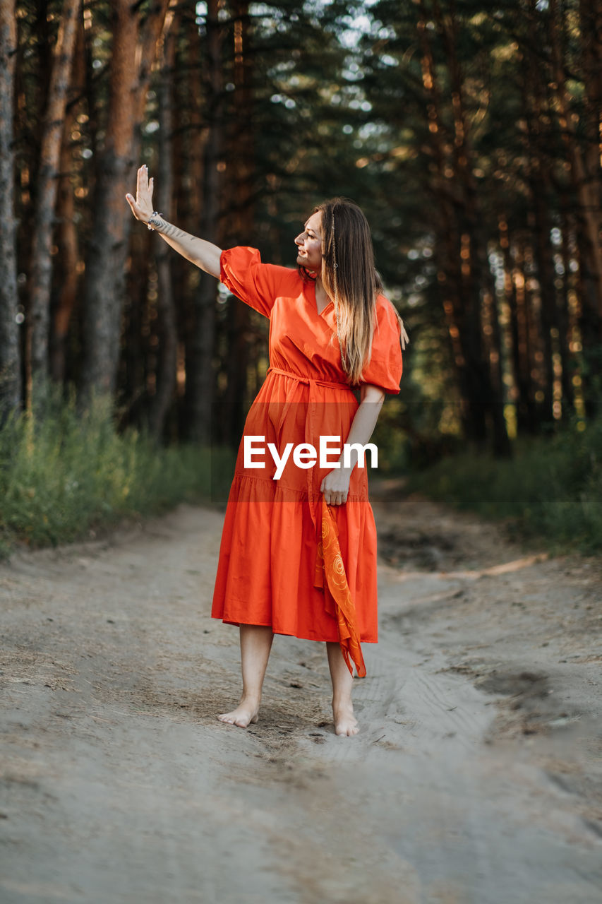 Barefoot happy young woman in red dress with hand raised dancing in pine forest at summer day.