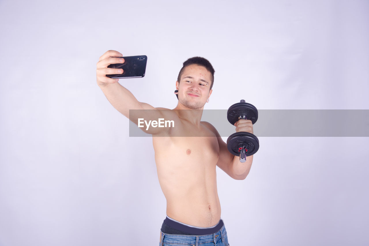 Shirtless man lifting dumbbell taking selfie with smart phone against purple background