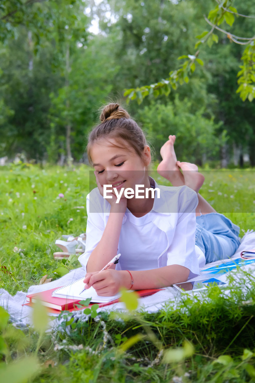 A young schoolgirl lies on the grass in the park with a notebook and a pen.