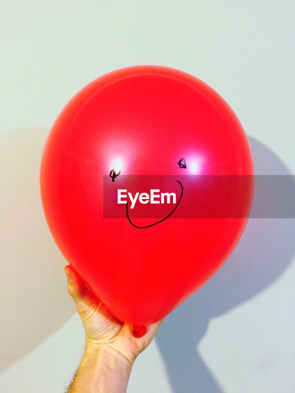 Cropped hand holding red balloon with smiley against white wall