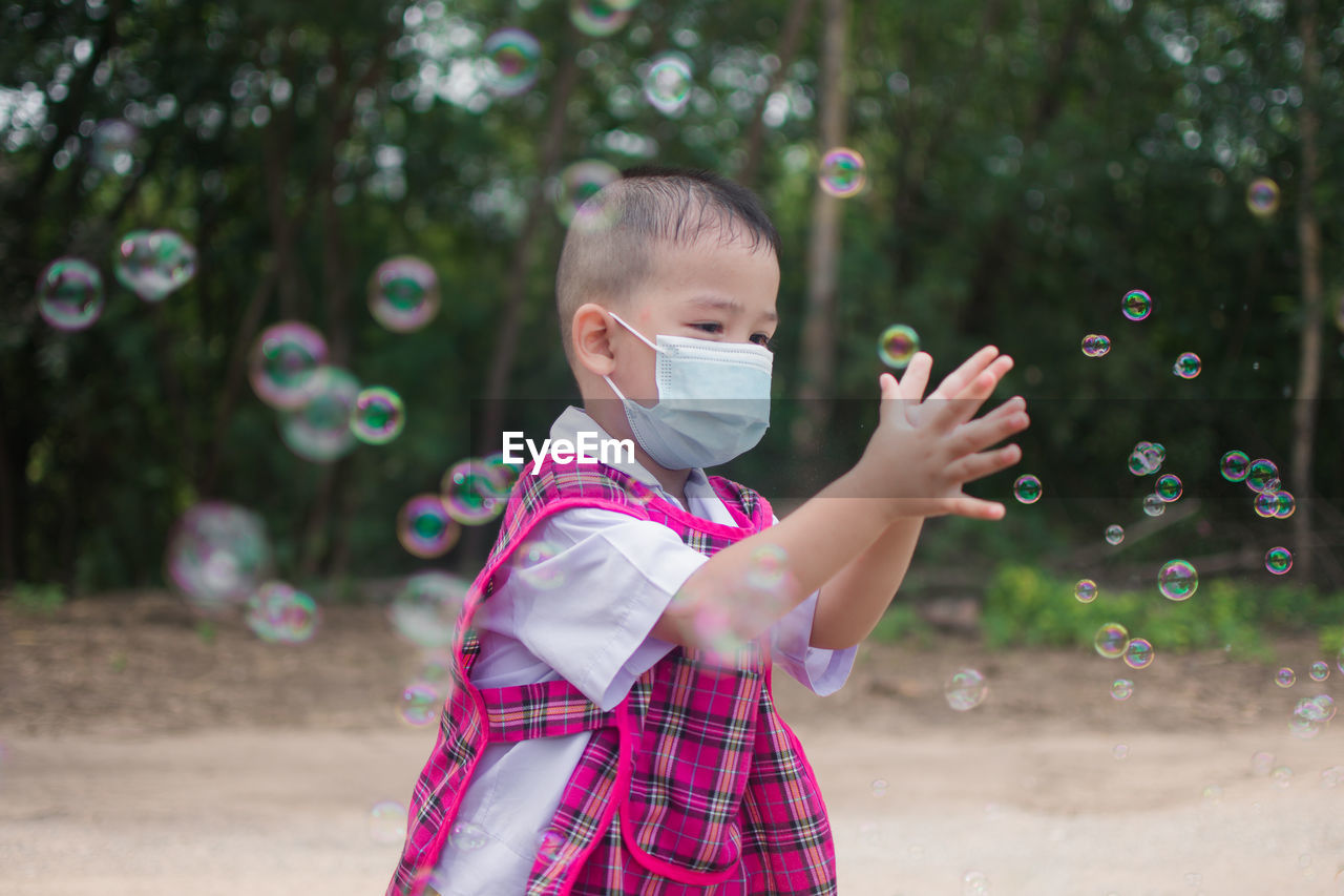 Boy wearing mask playing with soap bubbles standing outdoors