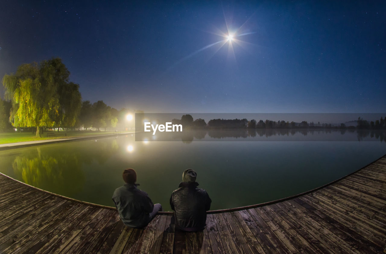 REAR VIEW OF TWO PEOPLE WALKING ON LAKE AT NIGHT