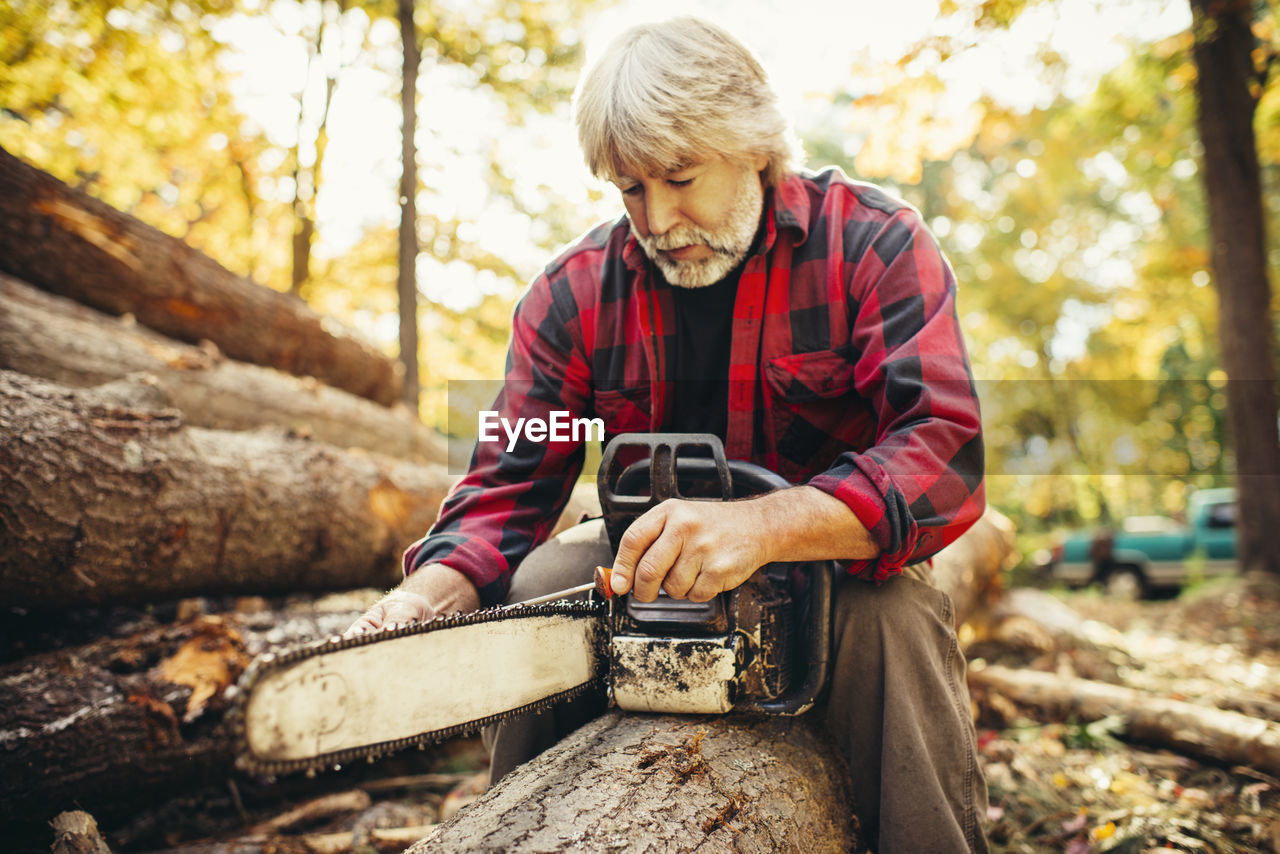 Male lumberjack examining chainsaw while sitting on log in forest