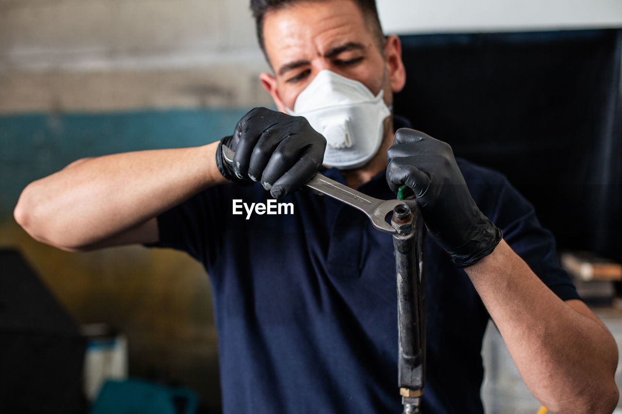 Male mechanic in respirator and latex gloves screwing nut on nozzle with spanner while working in professional workshop