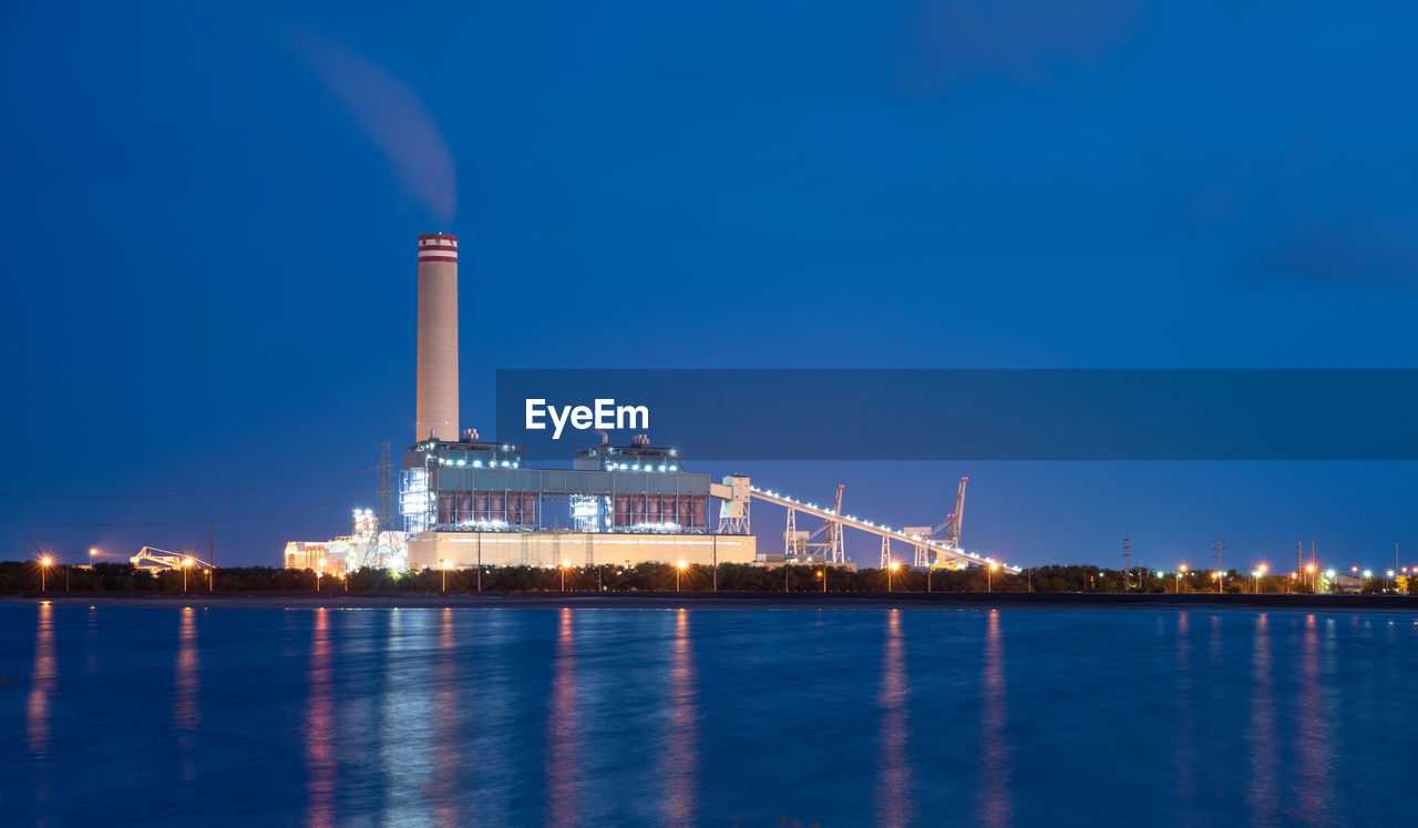 ILLUMINATED FACTORY BY SEA AGAINST CLEAR BLUE SKY