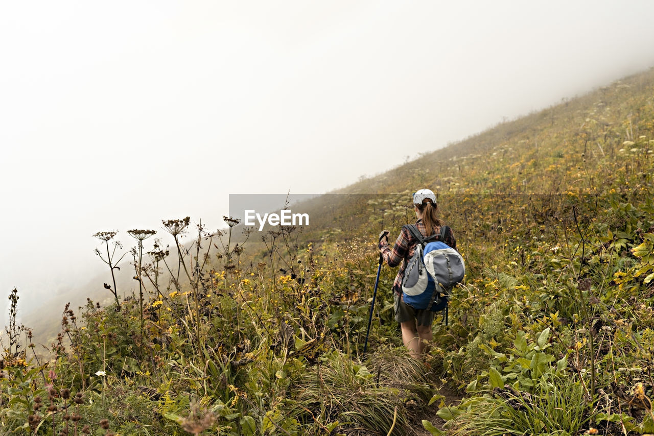 Rear view of young woman with big backpack trekking poles walking along mountain trail hiking in fog