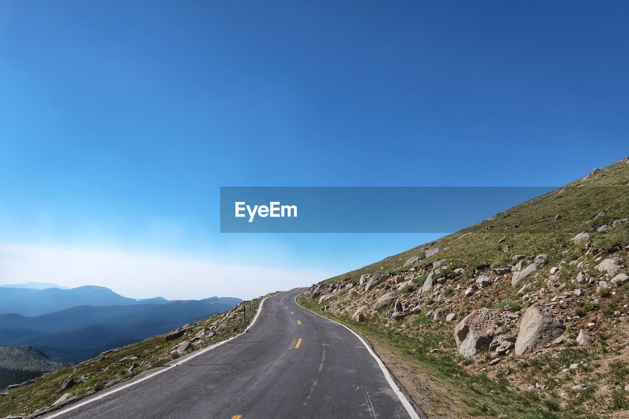 Road leading towards mountains against clear blue sky