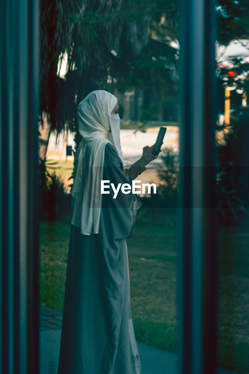 Reflection of a rear view of a muslim lady in a veil using phone on the window glass 