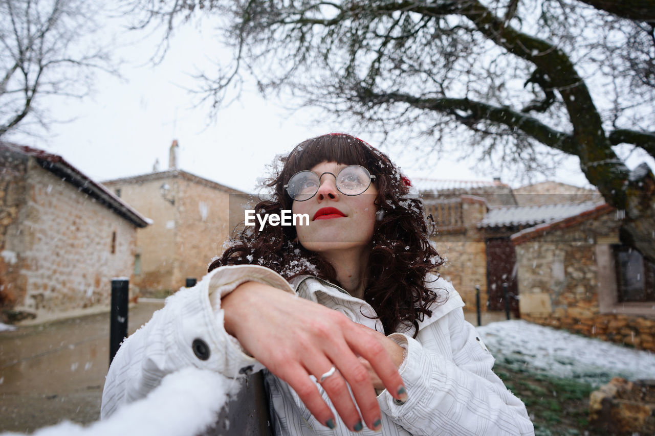 Young woman sitting on snow covered bench against sky