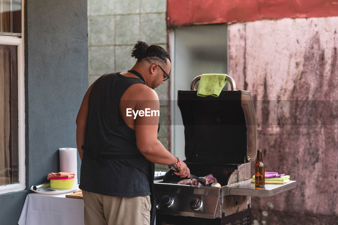 Back view of ethnic male in apron placing raw marinated meat on grill of barbecue cooker while cooking steaks