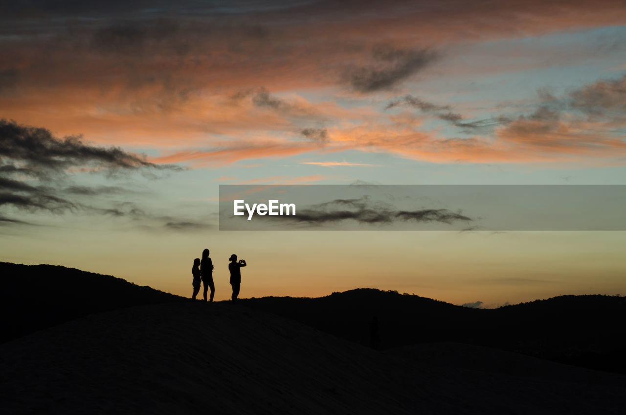 SILHOUETTE COUPLE STANDING ON LANDSCAPE AGAINST SKY AT SUNSET