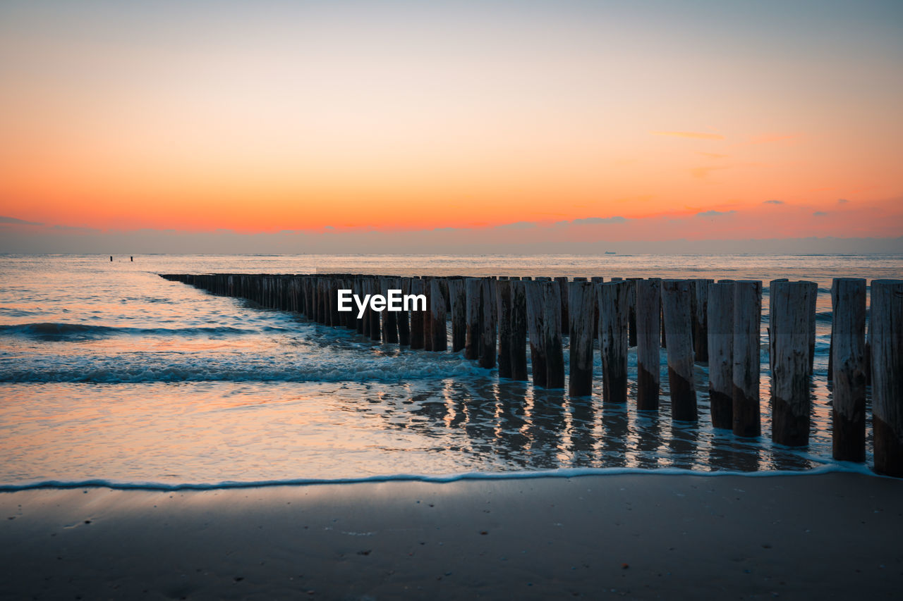 Wooden posts in sea against sky during sunset