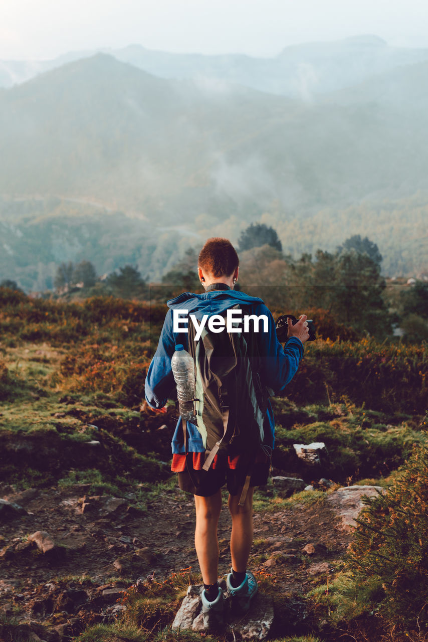 Back view of active touristic person in blue jacket and backpack climbing on ill with dry orange vegetation in spain