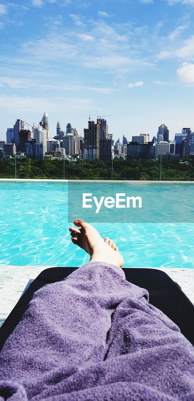Low section of person with purple towel relaxing by swimming pool in city during sunny day