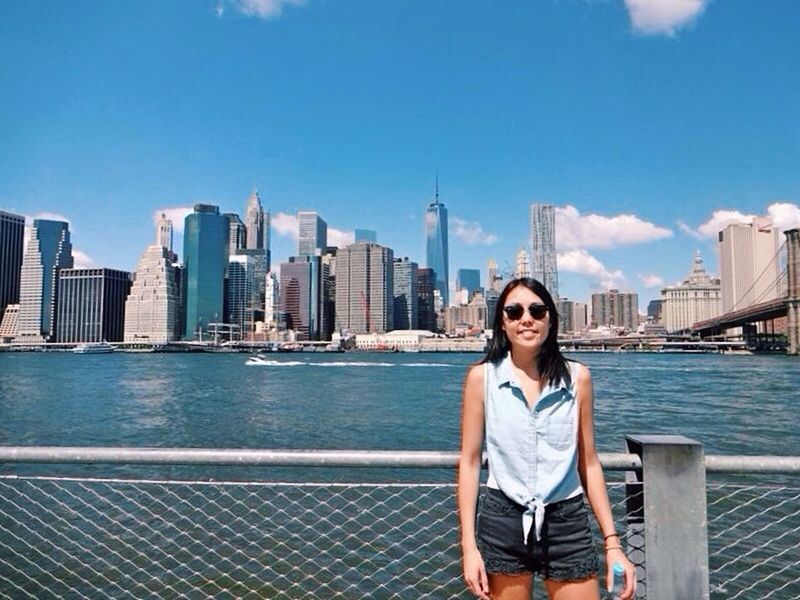 Portrait of young woman standing by railing and river against cityscape