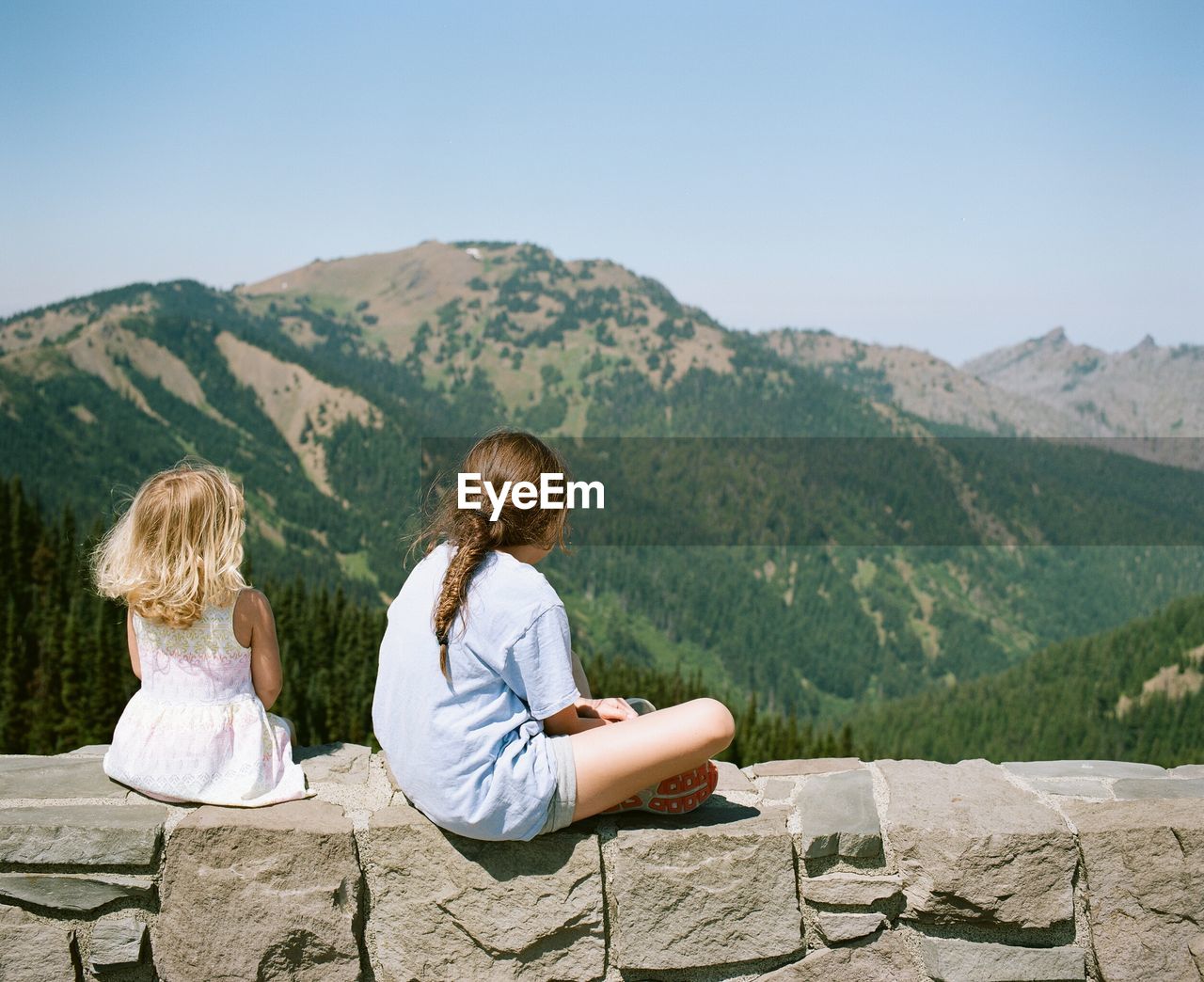 Rear view of girls sitting on stone wall against mountain