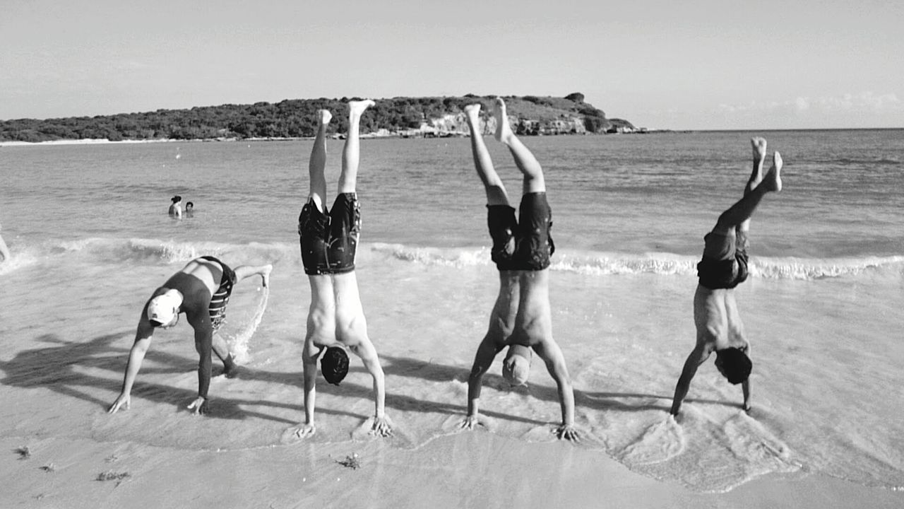 Upside down image of shirtless men performing handstand on beach against clear sky