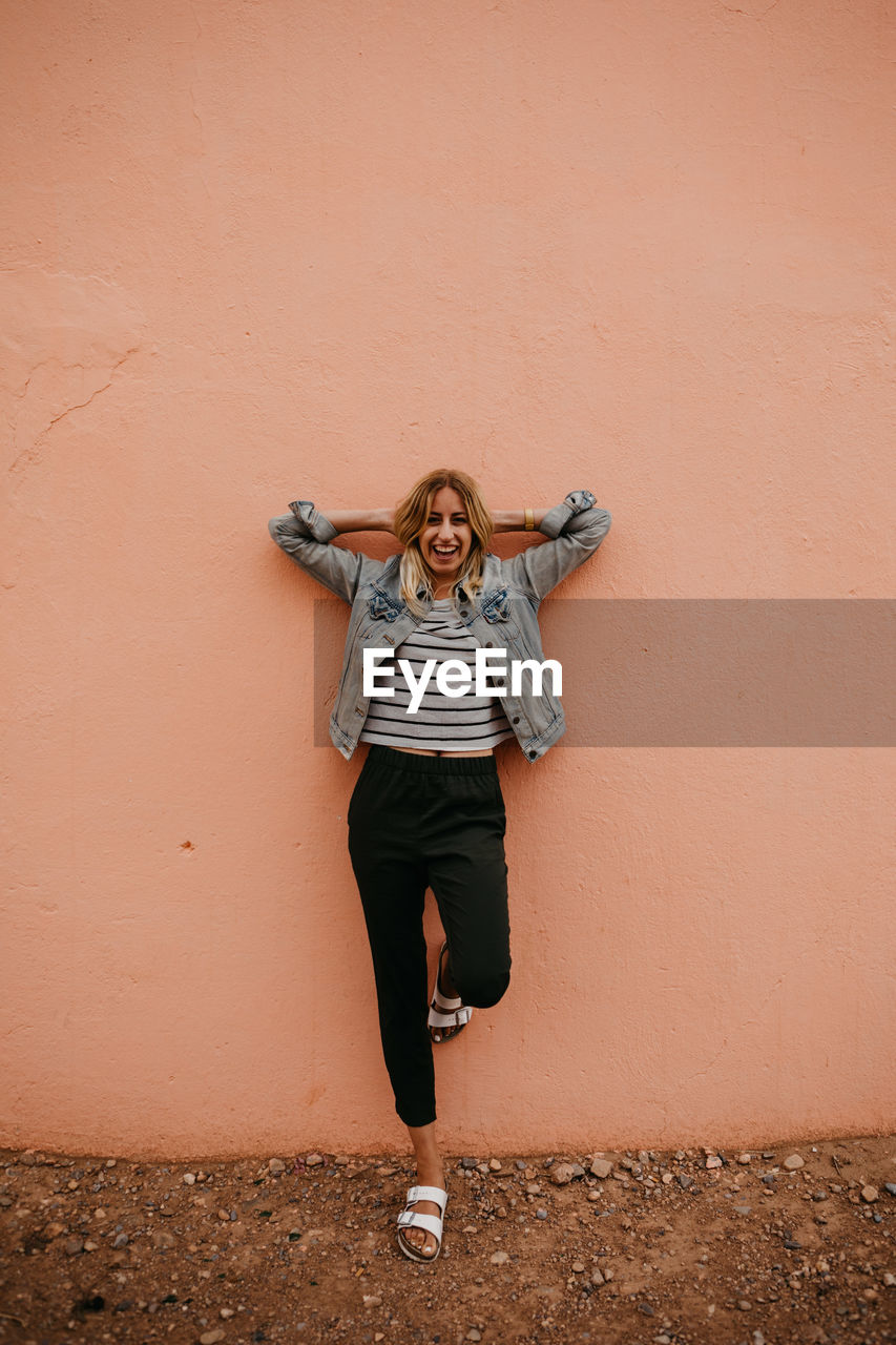 Portrait of happy young woman with hands behind head standing against wall