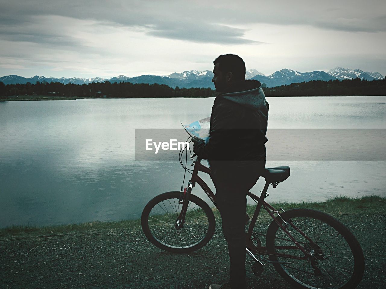 Man with bicycle and map by lake against sky