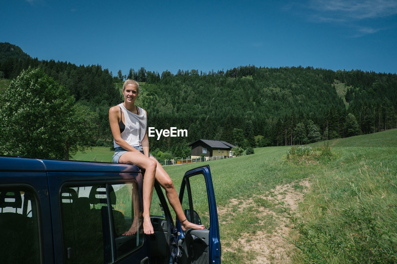 Portrait of smiling woman sitting on car roof against sky