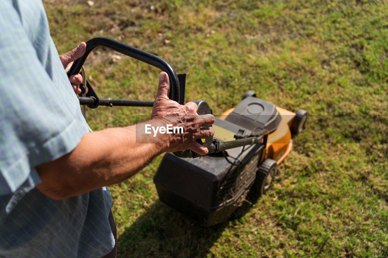 From above side view of male gardener mowing grassy lawn near bushes and trees in summer