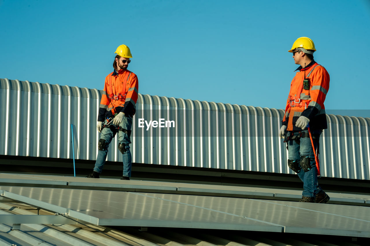 headwear, helmet, men, hard hat, architecture, occupation, adult, manual worker, industry, working, sky, hat, two people, protection, full length, built structure, person, standing, construction industry, construction worker, business, clothing, day, blue, cooperation, protective workwear, nature, reflective clothing, transportation, railing, outdoors, clear sky, teamwork, mature adult, roofer, communication, building exterior