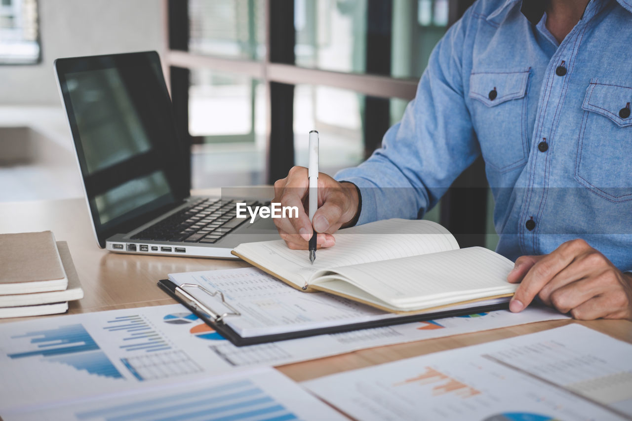 Midsection of businessman writing in book on table at office