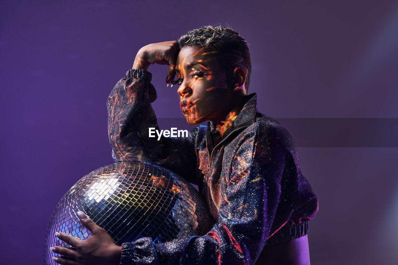 Side view of trendy young african american female with makeup looking at camera while holding glitter ball in violet light