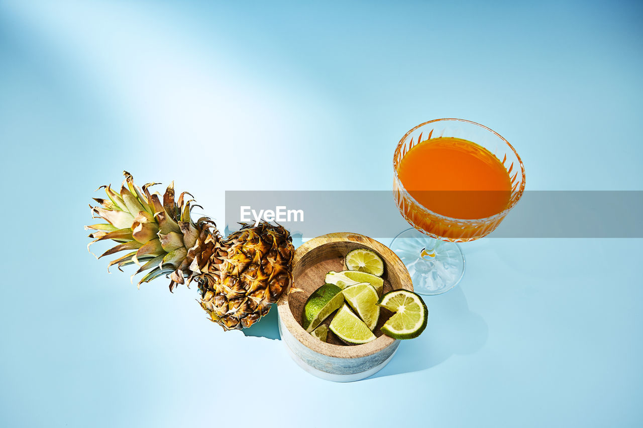 Directly above shot of orange drink on blue background with pineapple and lime garnishes
