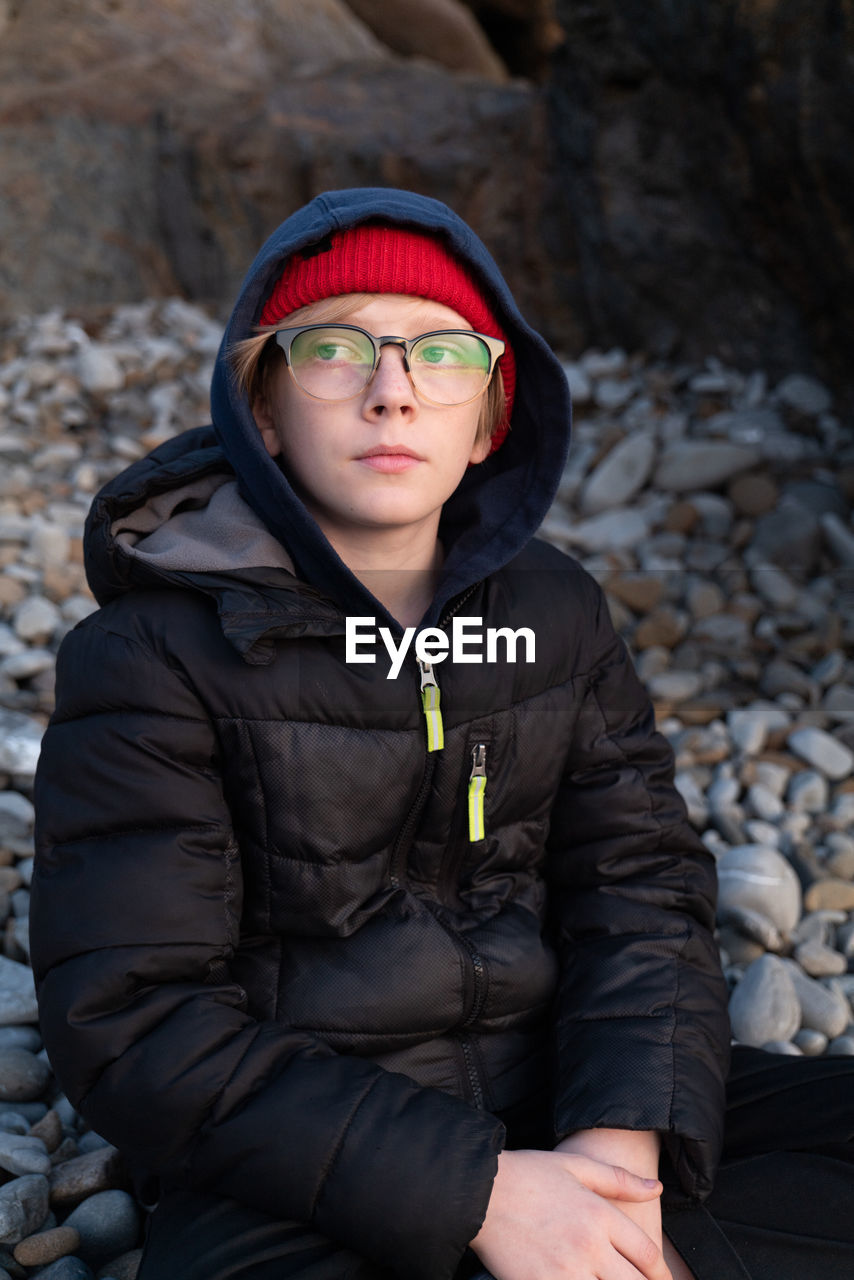 Tween boy sitting on beach of stones wearing glasses watching sunset