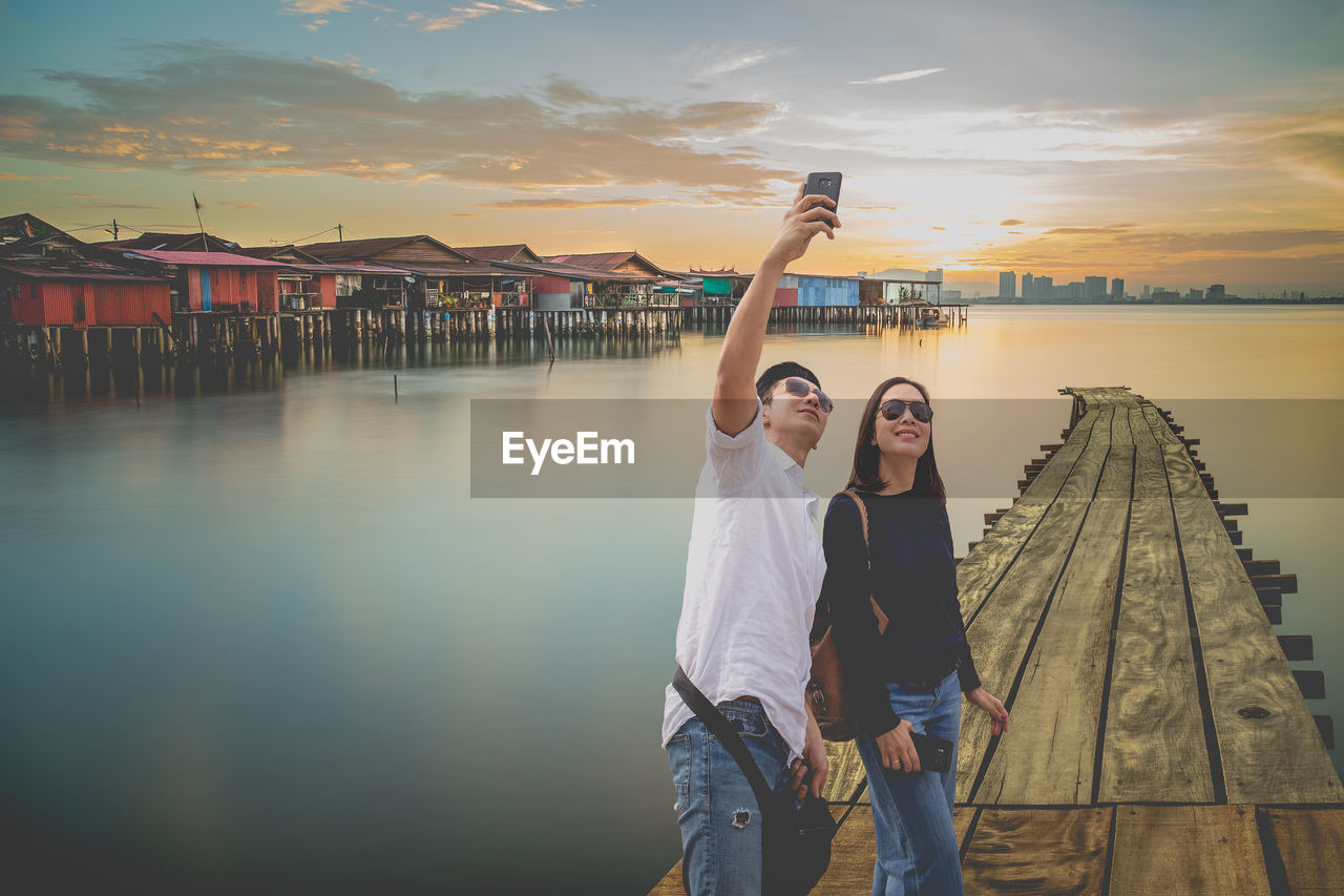 Man taking selfie with woman while standing on pier against sky during sunset