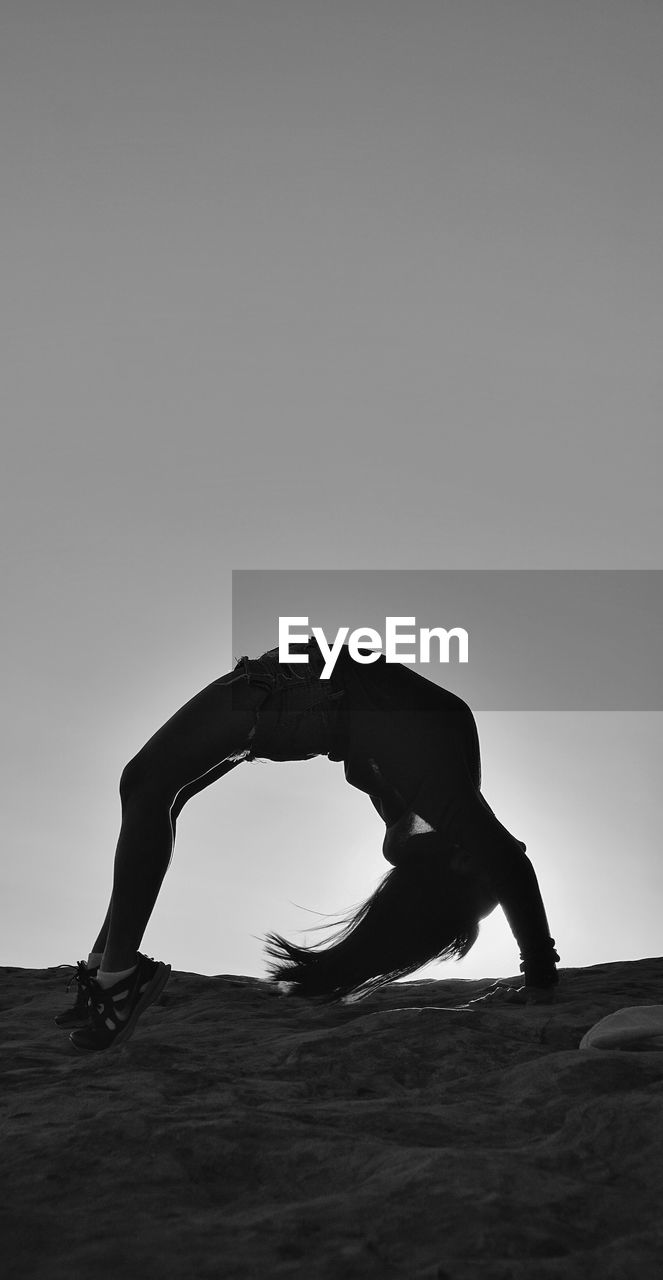 Side view of woman bending over backwards at beach against clear sky