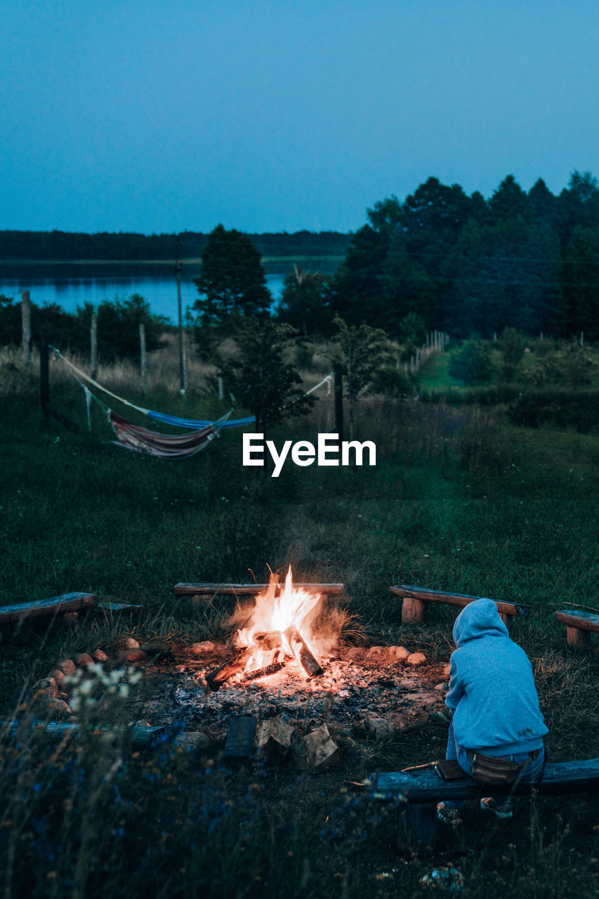 A person in a hood sitting next to the bonfire with hammocks, forest and a lake in the background.