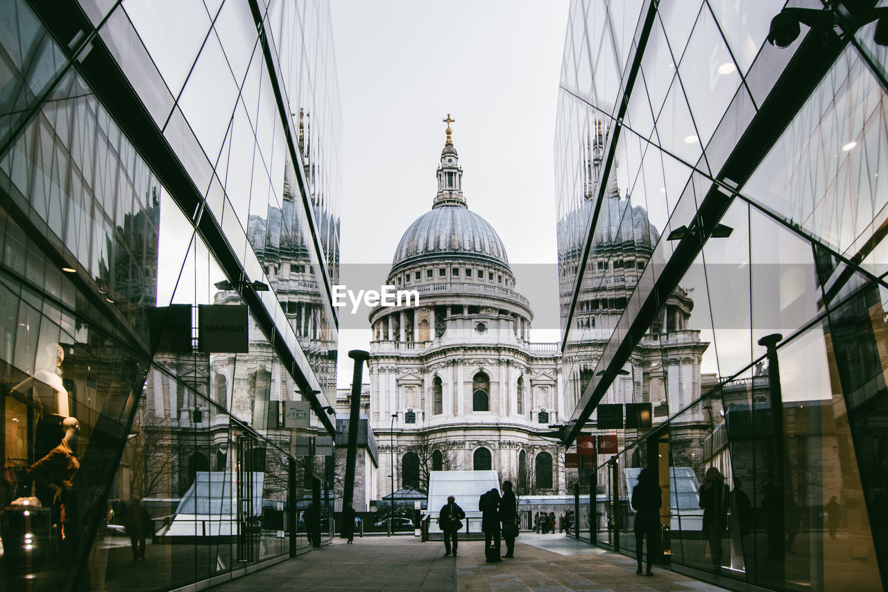 People on footpath amidst modern buildings by st paul cathedral against clear sky